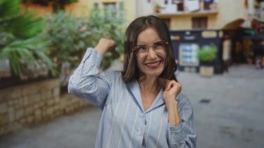 Gözlüklü kadın gülümsüyor ve parmaklarıyla güneşli bir cadde pazarında zafer işareti yapıyor; eğlence kutlaması seyahati.