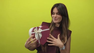 İspanyol kadın, kalp şeklinde hediye kutusunu sarı arka planda tutuyor neşeli bir ifade ve zarif bir saat sergiliyor..