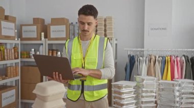 Yansıtıcı yelek giyen genç bir adam, kapalı bir odada dizüstü bilgisayar kullanırken kutularla ve kıyafetlerle çevrili bir hayır kurumunda bağış organize ediyor..