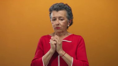 Kırmızı ceketli yaşlı kadın turuncu arka planda endişeli görünüyor. Endişelerini ve gerginliklerini elleri sımsıkı tutarak ifade ediyor..
