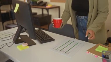 Modern ofiste çalışan İspanyol bir kadın, gözlük takıyor, bilgisayarda daktilo yazıyor, kahve fincanı tutuyor, çalışma ortamını somutlaştırıyor..