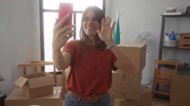 Yeni evinde cep telefonuyla gülümseyen İspanyol bir kadın. Parlak oturma odasındaki kutular ve mobilyalarla çevrili..
