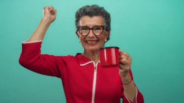 Kırmızı elbiseli, gri saçlı bir kadın elinde kırmızı bir kahve fincanı tutarken gülümsüyor, neşe ve neşe saçıyor..