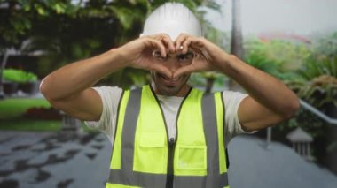 Beyaz kasketli ve neon renkli yelek giyen bir adam yeşil parkta gülümserken elleriyle kalbi şekillendirir..