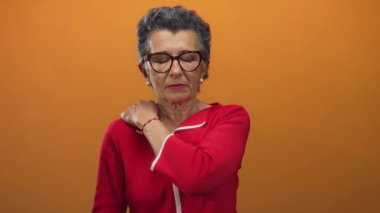 Kırmızı tişörtlü, gri saçlı ve gözlüklü yaşlı kadın turuncu arka planda omuz ağrısı yaşıyor, rahatsız edici ifadeleri ve yaşla ilgili sağlık sorunlarını vurguluyor..