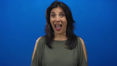 Orta yaşlı İspanyol bir kadın, parlak bir gülümseme ile mavi bir stüdyo duvarına karşı kendini işaret ediyor..