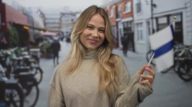 Meşgul şehir caddesinde elinde finiş bayrağı tutarken gülümseyen sarışın kadın; ulusal gurur vatanseverlik desteği.