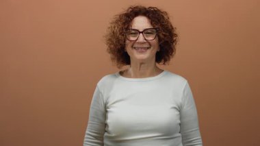 Gülümseyen ve kahverengi duvar arkasına el sallayan bir kadın, samimi ve cana yakın bir tavır sergiliyor..