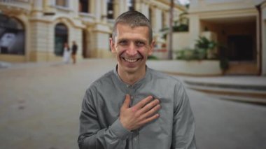 Açık havada gülümseyen adam, canlı bir şehir caddesinde duruyor, arka planda tarihi bir mimariyle pozitif bir hava sergiliyor..