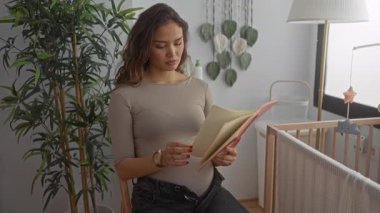 Sıcak bir yatak odasında beşiği ve dekoruyla kitap okuyan hamile bir kadın, genç yaşam ve aile hazırlıkları hakkında huzurlu bir anne atmosferi sunuyor..