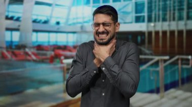 Kapalı bir teknede, büyük pencereleri ve şezlongları olan bir iç mekan ile çevrili, acı dolu bir ifadeyle boğazını sıkan bir genç adam..