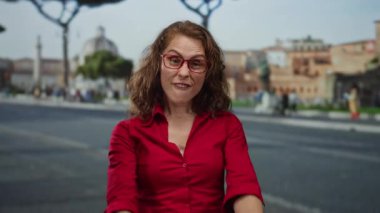 Kırmızı tişörtlü bir kadın, şehir sokaklarında tarihi binalarla anlamlı jestler yapıyor, açık havada duygusal bir tepki gösteriyor.