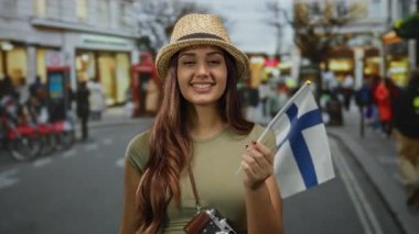 Elinde finnish bayrağı tutan kadın, arka planda sokak sahnesi olan, hasır şapka takan ve günlük giysiler giyen, canlı şehir keşiflerini yakalayan, gezgin neşesini vurgulayan.