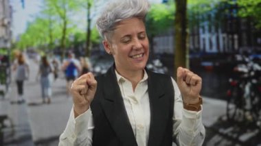 Kısa gri saçlı kadın, güneşli bir şehirde ağaçlarla ve park edilmiş bisikletlerle dolu bir sokakta yumruklarını kaldırıyor..