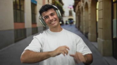 Kulaklıklı, beyaz gömlekli genç adam, bir şehir caddesinde gülümsüyor, müzikten hoşlanıyor ve ileriyi işaret ediyor, canlı bir şehir manzarası yakalıyor..
