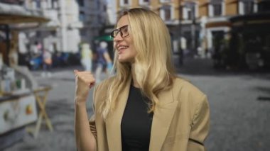 Kadın hareketli bir şehir caddesinde başparmağıyla mutlu bir şekilde el kol hareketi yapıyor, canlı bir gülümsemeyle gözlük takıyor, enerjik bir şehir manzarasını gün ışığında yakalıyor..