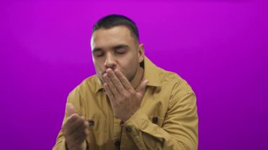 Kahverengi gömlekli genç İspanyol adam gözlerini kapatır ve eflatun stüdyo arkaplanına öpücük atar; şefkat sıcaklığı.