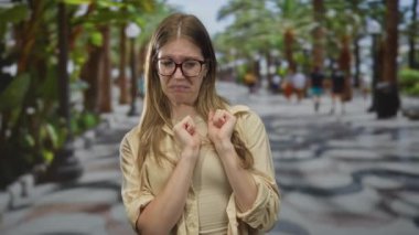 Gözlüklü genç sarışın kadın burnu kırışıyor ve güneşli yeşil bir parkta el kaldırıyor..