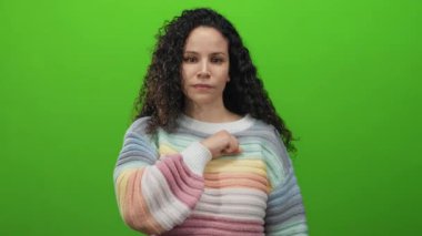 Kendinden emin genç kadın canlı yeşil duvara karşı yumruğunu uzatıyor. Kıvırcık saçı ve renkli kazağıyla güçlü ve kararlı..