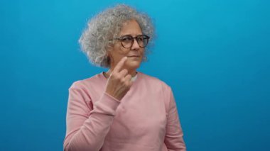 Gri saçlı ve gözlüklü yaşlı kadın, izole edilmiş mavi arka plana karşı düşünceli bir şekilde el kol hareketi yapıyor..
