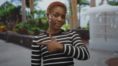 Genç Afro-Amerikalı kadın çizgili bir bluzla ağaçlı bir caddede baş parmağını kaldırdı..