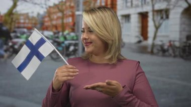 Sarışın kadın geniş bir şekilde gülümsüyor ve kalabalık şehir caddesinde el kaldırarak finnish bayrağını tutuyor; gurur.