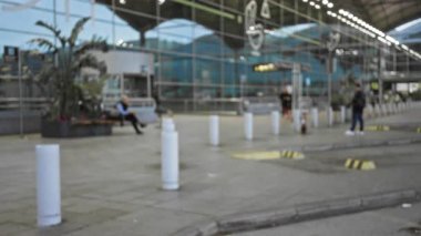 Havaalanı terminalinin dışındaki bulanık bir sahnede, arka planda, odakları kesilmiş mimari elementlerle çevrili insanlar ve bitkiler yoğun ama sakin bir atmosfer yaratıyorlar..