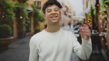 Genç İspanyol gözlüklü ve beyaz kazaklı adam restoran terasında kamerayı işaret ediyor..