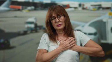 Üst düzey İspanyol kadın havaalanı terminalinde göğüs ağrısı yaşıyor. Arka planda uçaklar seyahat stresi ya da acil bir sağlık sorunu olduğunu gösteriyor.