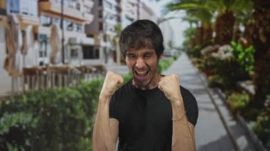 Güneşli bir sokakta neşeli bir sırıtışla yumruk kaldıran adam palmiye ağaçları, açık kafein oturma ve yeşil çalı kenarları ile kaplı;. 