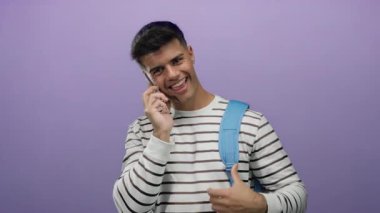 Genç adam gülümseyen akıllı telefon çizgili gömlek ve mavi sırt çantasıyla mor arka plana yaslanmış mutluluk ve pozitifliği gösteren başparmak işareti veriyor..