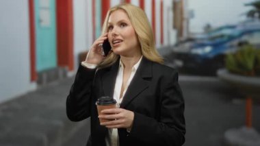 Açık havada akıllı telefondan konuşan bir kadın resmi takım elbise giyerken elinde kahve, arka planda şehir manzarası sarışın, genç ve profesyonel bir kadının modern şehir yaşam tarzını somutlaştırıyor..