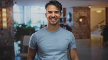 Mavi gömlekli genç adam modern otelin iç kısımlarında çiçeklerle ve parlak atmosferle öpüşürken neşeli bir ruh hali ve hoş geldin jestleri sergiliyor..