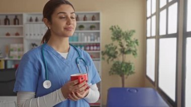 Mavi önlüklü genç bir hemşire arka planda bitki ve raflarla dolu parlak bir hastane odasında fincanı tutarak klinikte mola verilmesini öneriyor..