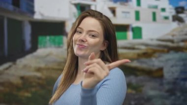 Uzun kahverengi saçlı beyaz bir kadın, güneşli sahilde, okyanus dalgalarıyla parmaklarıyla kalp hareketi yapar..