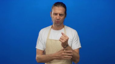 Uzun saçlı, beyaz tişörtlü, bej önlüklü sarışın adam sol bileğini kavrıyor ve mavi stüdyo ortamında irkilmeye başlıyor..