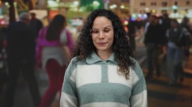 Kalabalık bir sokakta, gözlüklü ve süveterli bir şekilde gülümseyen bir kadın. Etrafı yürüyen insanlarla çevrili, canlı bir şehir atmosferi sağlıyor.