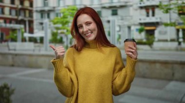 Şehir caddesinde kahveyle gülümseyen, sarı kazak giyen, baş parmak kaldıran, kızıl saçlı, ağaçlı ve mimarlı şehir manzaralı bir kadın..