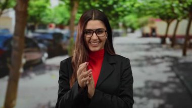 Açık bir sokakta, yeşil yapraklı bir tepe örtüsünün altında canlı bir şehir ortamında gülümseyen gözlük takan bir kadın..