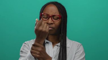 Gözlüklü bir kadın, yeşil bir duvara karşı bileklerini inceliyor. Odaklanmış bir ortamda tefekküre ya da endişeye işaret ediyor..