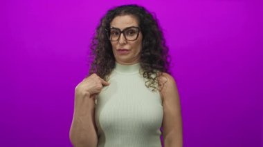 Kolsuz bluz ve gözlüklü İspanyol kadın mor bir stüdyoda kendini işaret ediyor..