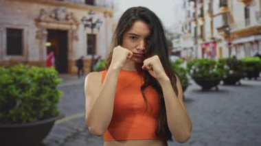 Turuncu kollu tişörtlü kadın, eski bina ve yeşilliğin önündeki kaldırım taşı sokakta yumruklarını sıkıyor; kararlılık dayanıklılığı cesareti.