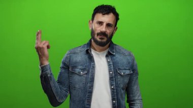 Sakallı İspanyol bir adam yeşil arka planda jest yapıyor, kot ceket ve beyaz gömlek giyiyor..
