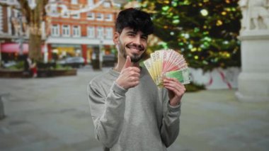 Dışarıda gülümseyen bir adam, elinde Norveç kron banknotları arka planda festival ışıkları ve binaları olan bir şehir sokağında başparmak kaldırıyor..