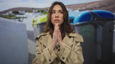 Şehir sokaklarında renkli çöp tenekeleri ve arka planda binaları olan açık havada dua eden kadın şehir ortamında umut dolu ve huzurlu görünüyor..