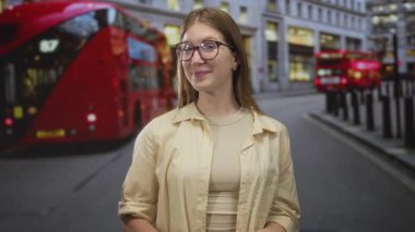 Yoğun bir şehir caddesinde kırmızı bir çift katlı otobüsün yanında gülümseyen gözlüklü genç sarışın kadın, iyimserlik..