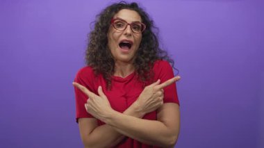 Ağzı açıkken mor stüdyoda kıvırcık saçlı, işaret eden ve kollarını kavuşturan gözlüklü bir kadın..