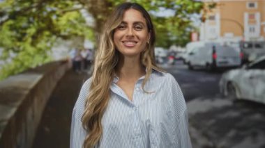 Genç sarışın kadın yüzü, çizgili bluz içinde dalgalı saçlarıyla gülümsüyor yapraklı güneş ışığı kaldırımında; keyif güveni.