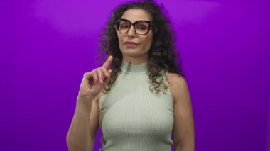 Kıvırcık saçlı, büyük gözlükler takan ve mor stüdyo ortamında kolsuz, yeşil bir üst parmağı olan bir kadın..