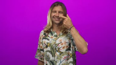Tropikal tişörtlü, uzun sarı saçlı adam mor stüdyo setinde parmaklarını burun buruna gösteriyor..